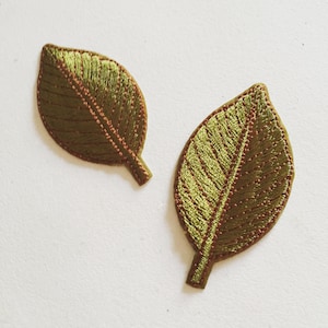 May include: Two green embroidered fabric leaves with brown stitching. The leaves are on a white background.