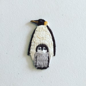May include: Embroidered patch featuring a white and black emperor penguin with a smaller grey and black penguin chick inside. The patch is ideal for adding a touch of whimsy to clothing, bags, or other accessories.