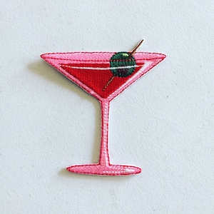 Pode incluir: Copo de martini bordado rosa com uma azeitona verde em um palito.