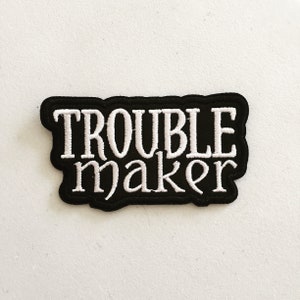 Pode incluir: Etiqueta bordada preta com letras brancas que dizem "Trouble maker".