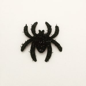 Könnte beinhalten: Eine schwarze Spinnendekoration mit acht Beinen und einer strukturierten, glitzernden Oberfläche. Die Spinne ist eine Dekoration zum Thema Halloween, ideal, um jedem Raum eine gruselige Note zu verleihen. Die Spinne ist vor einem weißen Hintergrund.