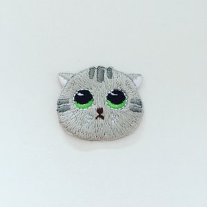 May include: Embroidered grey cat patch with green eyes and a brown nose. The cat has a sad expression.