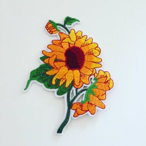 May include: Embroidered iron-on patch featuring a yellow sunflower with a red centre and green leaves. The sunflower is surrounded by two smaller yellow flowers.