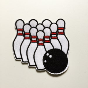 Pode incluir: Um conjunto de pinos de boliche brancos e vermelhos com uma bola de boliche preta. Os pinos estão dispostos em forma de triângulo com a bola na frente.