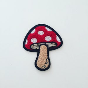 Könnte beinhalten: Gestickter Pilzaufnäher mit rotem Hut und weißen Punkten. Der Pilz hat einen beigefarbenen Stiel und einen schwarzen Rand. Ideal zum Aufnähen auf Kleidung oder Accessoires.