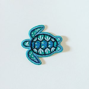Op de afbeelding: Een geborduurde strijk-op patch van een blauwe en groene zeeschildpad met een geometrisch patroon op zijn schild.