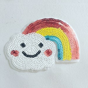 Könnte beinhalten: Eine weiße Wolke mit einem lächelnden Gesicht und rosa Wangen, hinter der sich ein Regenbogen aus Pailletten befindet. Der Regenbogen ist rot, orange, gelb, grün, blau und lila.
