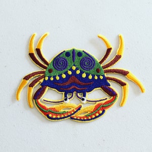 May include: A colourful embroidered patch of a crab with yellow, green, blue and purple details. The crab has a detailed shell with swirls and dots.