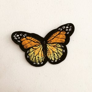 May include: A black and orange embroidered butterfly patch. The butterfly has black outlines and yellow accents on its wings.