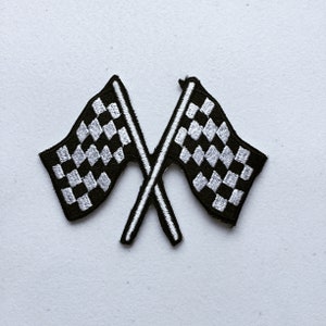May include: Two embroidered black and white chequered racing flags crossed over each other. The flags have a black background with white chequered patterns and white borders. These are likely patches for clothing or accessories.