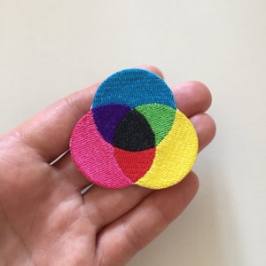 Op de afbeelding: Een kleurrijk geborduurd patch met een ontwerp van overlappende cirkels in tinten blauw, groen, paars, rood, zwart en geel.