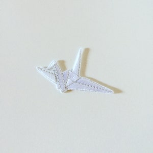 Op de afbeelding: Witte geborduurde origami-kraan-patch. De kraan is gevouwen in een traditionele stijl en heeft een delicaat, gedetailleerd ontwerp.