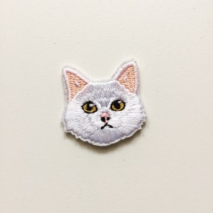 May include: Embroidered patch featuring a cat's head with a sad expression. The cat has gray fur, yellow eyes, and pink inner ears and nose. The patch is likely designed to be ironed or sewn onto fabric.