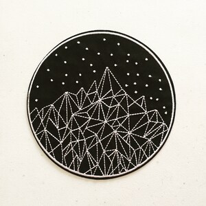May include: A circular, black embroidered patch with a mountain range design. White thread is used to outline the geometric peaks and a starry sky. The patch has a white border.