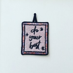 May include: A pink and white embroidered patch with black stitching. The patch is rectangular and has a black border. The text on the patch reads "do your best" with a star on either side of the text.