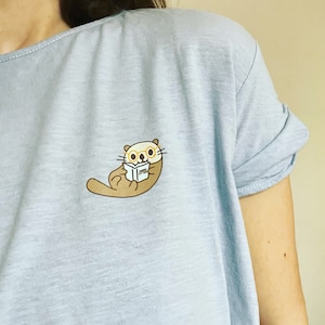 May include: A light blue t-shirt featuring a cartoon illustration of an otter wearing glasses and reading a book that says "Happy Otter".