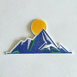 Può includere: Una toppa ricamata che rappresenta una catena montuosa blu e bianca con un sole giallo sopra. La catena montuosa ha accenti verdi.