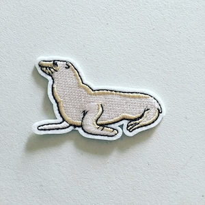 May include: An embroidered beige sea lion patch with white stitching around the edges. The patch is on a white background.