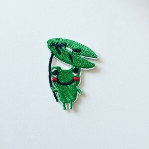 May include: Green embroidered frog with a big smile holding a green leaf over its head. The frog has black eyes and red cheeks.
