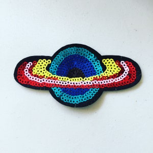 May include: A planet-shaped patch with a black felt backing. The planet is decorated with blue, yellow, red, pink, and white sequins. This decorative patch could be used to embellish clothing or accessories.