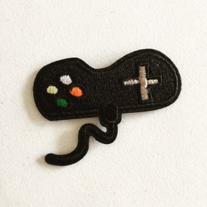 Könnte beinhalten: Schwarzer gestickter Patch mit einem Videospiel-Controller mit schwarzem Kabel. Der Controller hat bunte Knöpfe und ein weißes Pluszeichen.