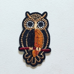 May include: A black and gold embroidered owl with orange sequins on its chest, perched on a branch. The owl has large, white eyes and a detailed design.