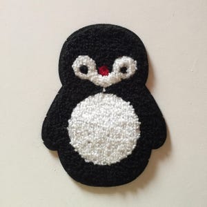 May include: A black and white penguin-shaped patch with a textured, tufted surface. The design features a large white circular belly, white eye patches, and a small red beak. The edges are defined by the black tufted material in a rounded shape.