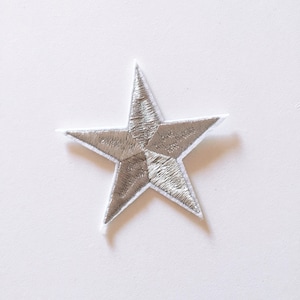 May include: A five-pointed star patch featuring metallic silver embroidery thread with a distinct sheen. The star has a white fabric border outlining its shape, creating a sharp contrast against the reflective silver centre of the embroidered design.
