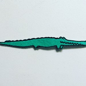 May include: A green alligator patch with a black outline. The patch features a stylized alligator design, with a long body and a detailed head. It is likely intended for clothing or accessories.