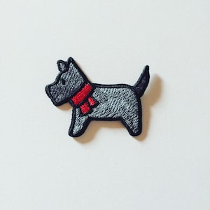 May include: Embroidered patch of a grey dog wearing a red scarf. The dog has a black outline and a textured appearance. The patch is designed for application on clothing or accessories, offering a decorative element.