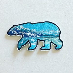 Può includere: Una toppa ricamata di un orso blu con una scena di montagna al suo interno. L'orso è contornato di nero e la scena di montagna è di un blu chiaro con cime bianche.