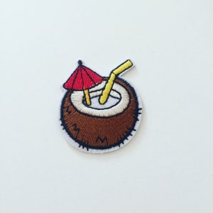 Pode incluir: Etiqueta bordada de um coco castanho com um canudo amarelo e um guarda-chuva vermelho. O coco está cheio de líquido branco.