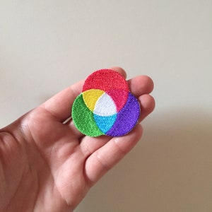 Op de afbeelding: Een kleurrijk geborduurd patch met drie overlappende cirkels in rood, geel, blauw, groen en paars.