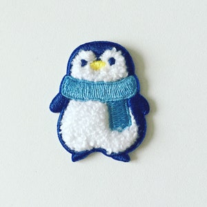 May include: Blue and white embroidered penguin patch with a blue scarf. The penguin has a yellow beak and black eyes.