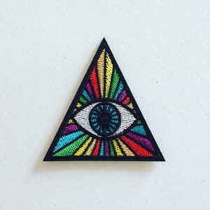 Könnte beinhalten: Ein farbenfroher gestickter Patch in Form eines Dreiecks mit einem großen Auge in der Mitte. Das Auge ist weiß mit einer blauen Iris und einer schwarzen Pupille. Das Dreieck ist mit regenbogenfarbenen Strahlen gefüllt, die vom Auge ausgehen.