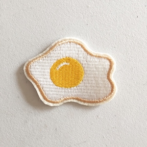 May include: A white embroidered patch shaped like a fried egg with a yellow yolk.