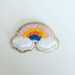 Könnte beinhalten: Ein regenbogenfarbener Filz-Patch mit goldener Glitzerbordüre. Der Regenbogen hat rosa, gelbe, orange und blaue Streifen. Der Patch ist in Form einer Wolke.