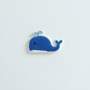 May include: A blue whale applique with white stitching and a black eye. The whale is embroidered on a white background.