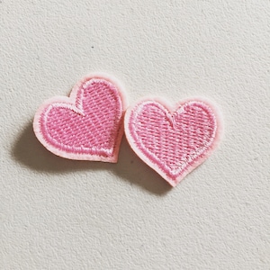 May include: Two embroidered heart patches. The hearts are a vibrant pink colour with a lighter pink border. The stitching detail is visible, creating a textured appearance. These patches are ideal for adding a touch of love to clothing or accessories.
