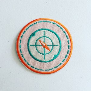 Op de afbeelding: Een ronde, oranje en roze geborduurde patch met een wit kompasontwerp erin. Het kompass heeft een groene omtrek en een oranje naald.