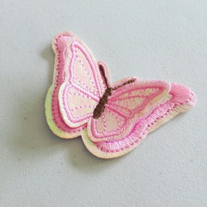 May include: A pink iridescent butterfly embroidered patch with brown accents. The patch is made of fabric and has a stitched edge.