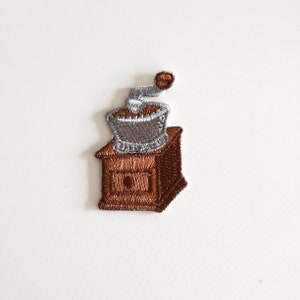 Könnte beinhalten: Gestickter Aufnäher mit einem Kaffeemühlen-Design. Der Aufnäher zeigt einen braunen Holzsockel mit einer Schublade, gekrönt von einer silbernen und braunen Mühle. Der Griff ist braun, und das Design ist detailliert mit verschiedenen Garnfarben.