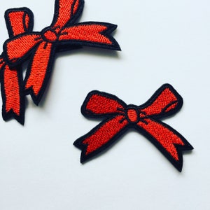 Könnte beinhalten: Zwei rote Stoffschleifen mit schwarzen Umrissen. Die Schleifen sind gestickt und haben ein einfaches Design.