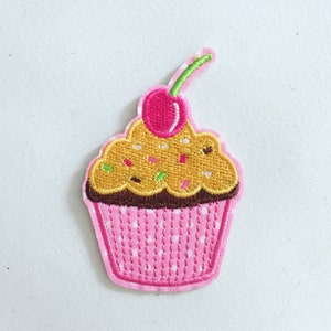 May include: A pink cupcake with brown icing and yellow sprinkles topped with a green stalk and a pink cherry. The cupcake is embroidered on a white fabric.