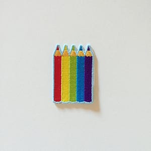 May include: A rainbow colored embroidered patch of five pencils. The pencils are red, orange, yellow, blue, and purple.