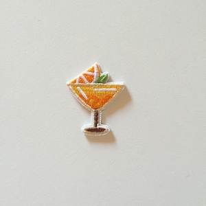 Op de afbeelding: Een geborduurde strijk-op patch van een geel cocktailglas met een sinaasappelschijfje en een groen blaadje erop.