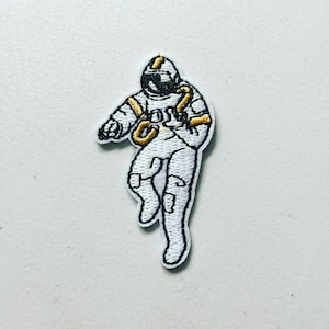 May include: White embroidered patch of an astronaut in a space suit with gold accents. The astronaut is floating in space with one arm raised.