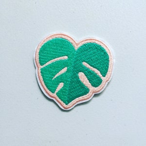 May include: Embroidered patch with a green monstera leaf design, a light pink border, and white backing. The heart-shaped patch has a detailed, textured appearance, suitable for clothing or accessories.