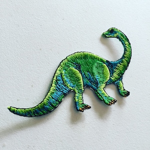 Pode incluir: Patch de dinossauro bordado em verde e azul. O dinossauro está em pé sobre suas quatro patas com a cabeça levantada e o pescoço arqueado.