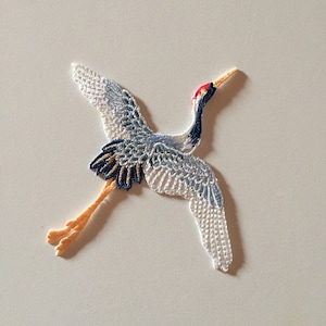 May include: A white, blue, and orange embroidered crane applique. The crane is in flight with its wings spread wide. The crane has a red beak and a long neck.